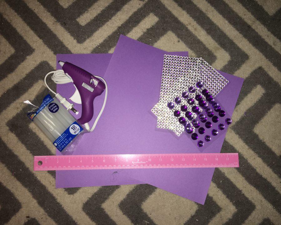 A collection of materials for designing a graduation cap: a hot glue gun and sticks, purple paper, a ruler and jeweled stickers.