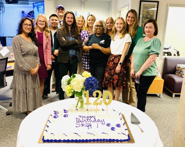 A group of people pose together behind a table that holds a vase of yellow and blue hydrangeas and a white sheet cake with "Happy birthday Skiff" written in purple and 120 in gold numbers stuck into it. 