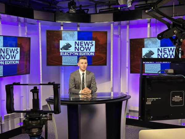 A news anchor, seated from behind the news desk, reads from the teleprompter. The screens behind him show a graphic in red, white and blue that reads "News Now Election Edition."